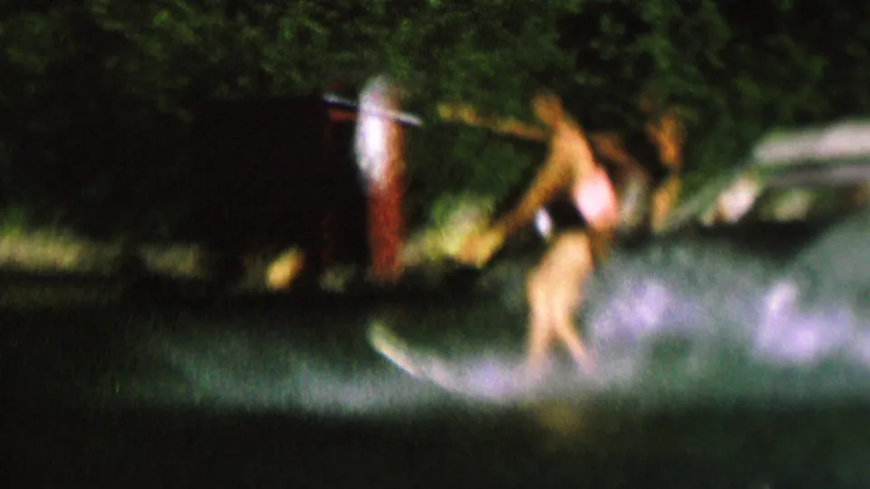 1962 8mm frame showing a water skier in a swimsuit cutting across the lake with trees and a shoreline behind them.