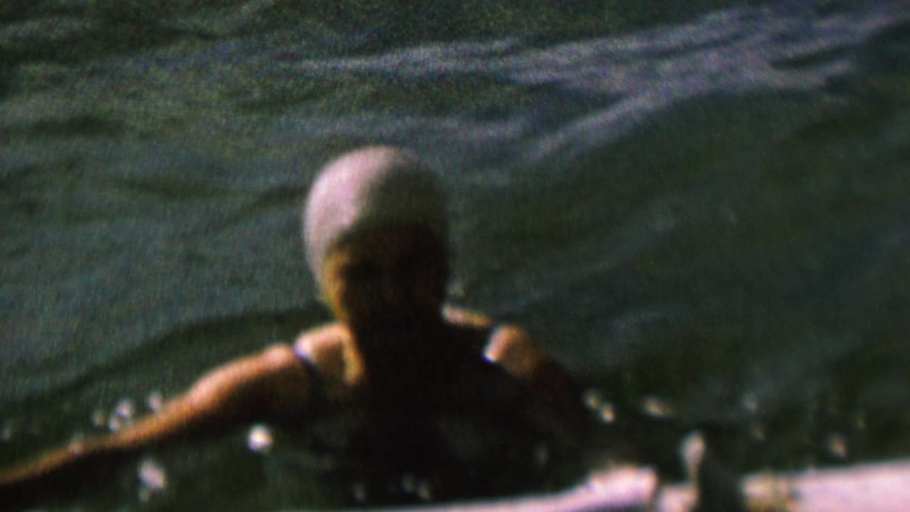A swimmer wearing a light cap and swimsuit is seen close to the camera in rippling lake water, with soft 8mm focus and heavy shadow on the face.