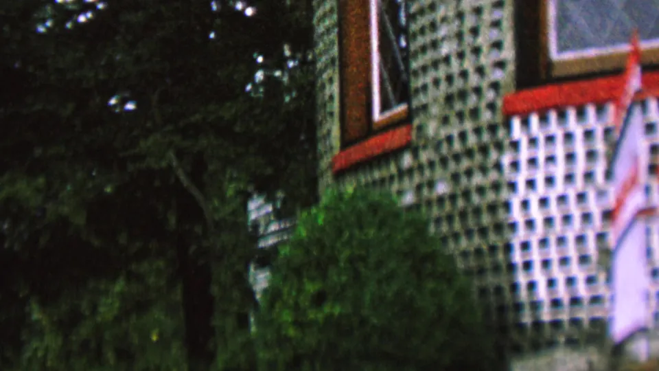 A closer home-movie view of the handmade glass-bottle wall shows red-trimmed windows, a concrete lattice surface, shrubs, and a small flag-like cloth in front.