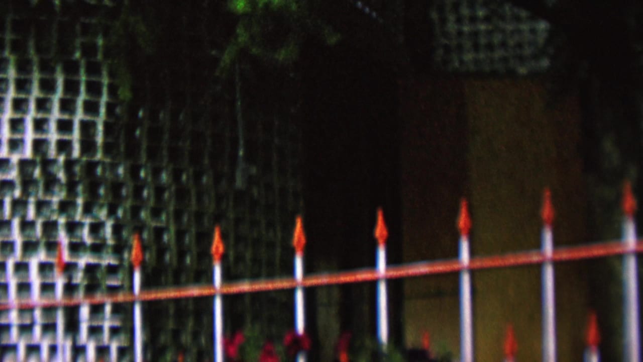 1962 St. Paul home-movie frame showing white vertical fence posts with red rails and pointed finials before a shaded green lattice wall and trees.