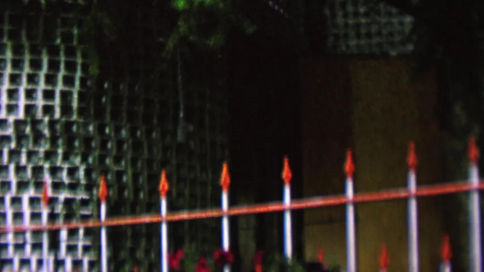 1962 St. Paul home-movie frame showing white vertical fence posts with red rails and pointed finials before a shaded green lattice wall and trees.