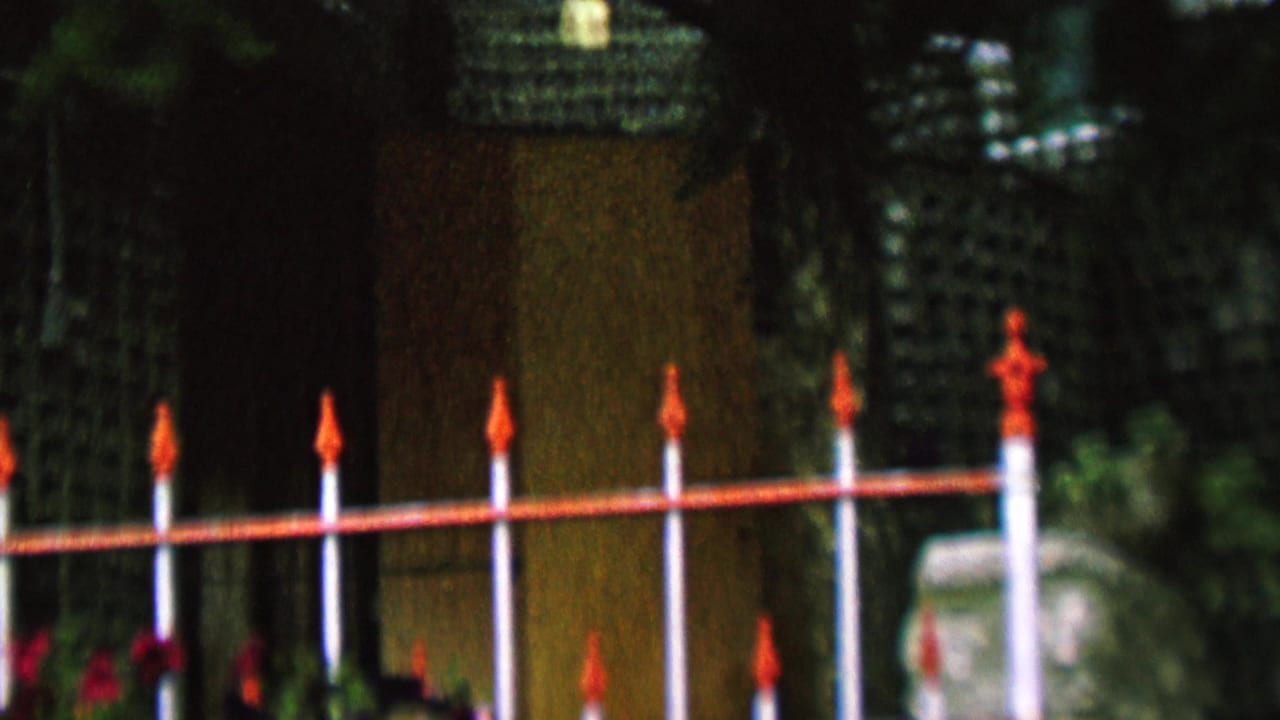Red and white fence continues across a lattice wall with foliage and a pale stone marker at right.