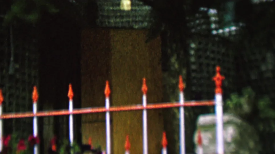 1962 St. Paul frame with the red-railed white fence crossing in front of a textured green lattice wall, foliage, and a pale stone marker near the right edge.