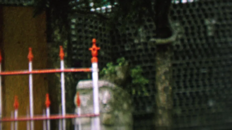 1962 St. Paul home-movie frame showing a closer section of the ornamental red and white fence with green gridded wall texture and a pale stone marker in the background.