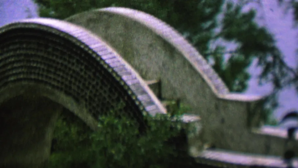 Side angle of the curved footbridge-like structure, with mosaic-like stone texture, leafy plants, and soft mid-century home-movie color.