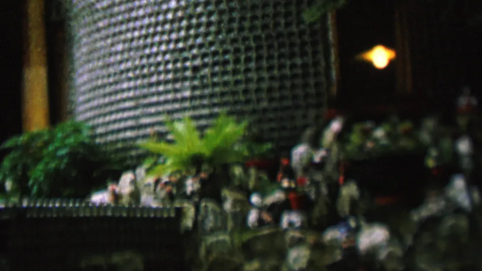 Soft 8mm frame of a nighttime St Paul exterior in 1962, with warm light inside a dark doorway and plants set along a stone-like ledge beneath a textured wall.