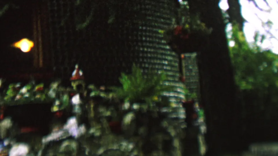 1962 St. Paul home-movie frame showing the central portion of a decorative outdoor installation, with figurines arranged near plants beneath a glowing light and textured bottle wall.