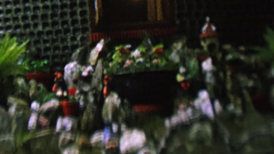 This 8mm frame presents a closer view of the miniature display, with painted rocks, small human-like figures, red planters, green plants, and dark recessed areas behind them.