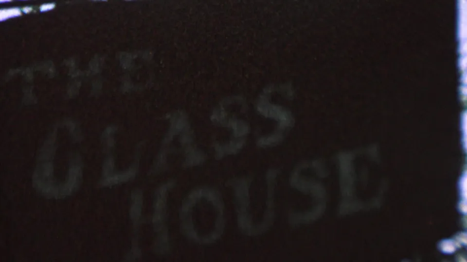 Shadowed 8mm view of the words The Glass House on a worn dark sign, with soft focus and grain obscuring the edges.