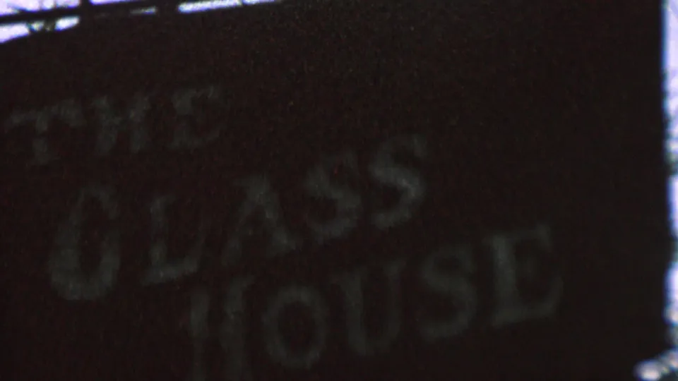 Low-light 1962 home-movie frame with the Glass House sign partly cropped and tilted, its white letters blurred by grain and motion.