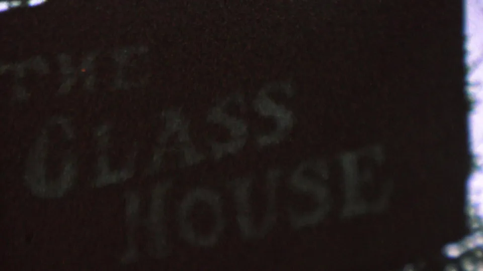 Final 8mm frame of the dark Glass House sign in St. Paul, with faded lettering, rough texture, and bright edge artifacts from the film scan.