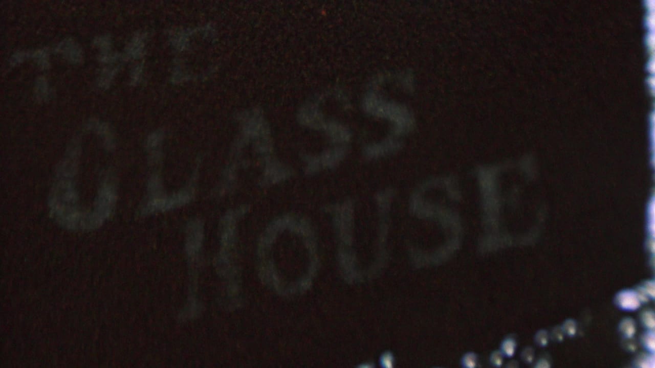 Tight 1962 St. Paul 8mm frame showing the dark textured sign for The Glass House, with pale lettering partially softened by grain and focus.