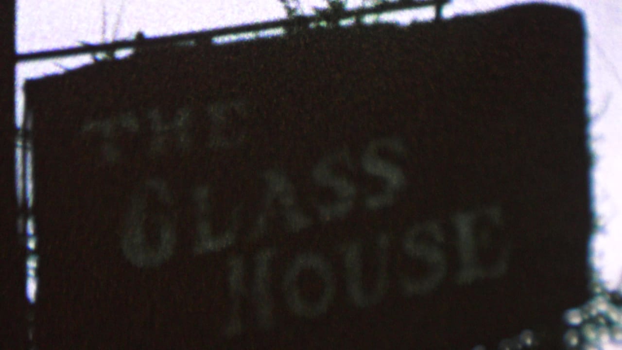 Dark sign with The Glass House lettering against a pale sky and overhead wires.