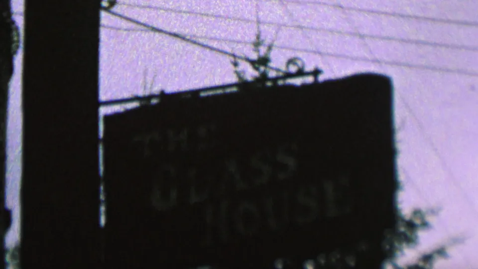 Wider 1962 St. Paul 8mm view of The Glass House sign mounted near a pole, with utility lines, pale sky, and tree branches visible around it.