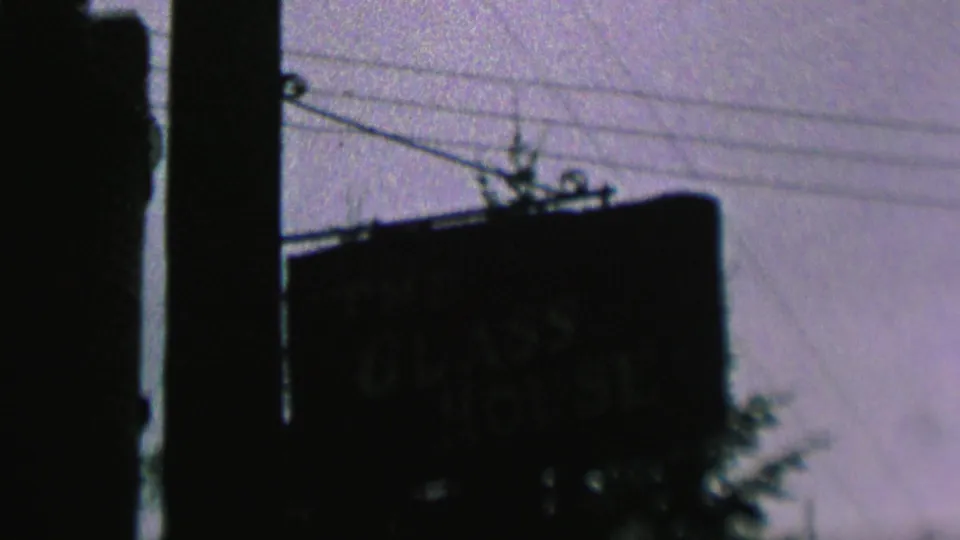 End frame from the 1962 St. Paul clip showing The Glass House sign from farther back, framed by a pole, overhead wires, pale sky, and foliage.