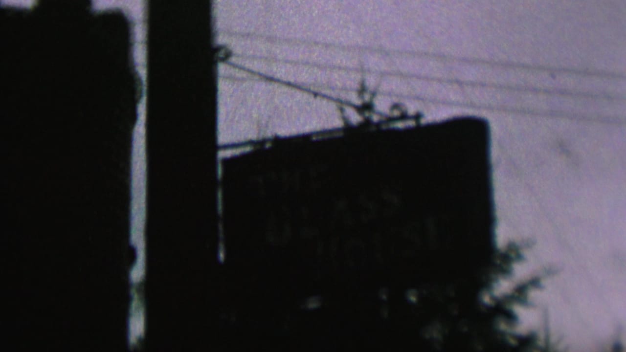 Low-light 1962 St. Paul home-movie frame showing a dark sign silhouette near utility poles, overhead wires, trees, and a purple-tinged sky.