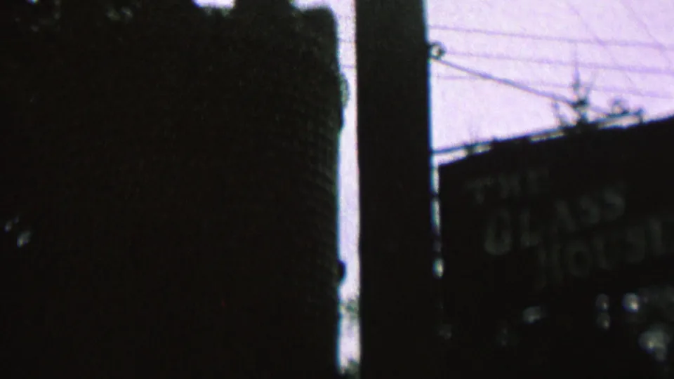 Frame from the St. Paul 8mm clip with a dark vertical structure dominating the view, a partially visible sign, utility lines, and soft tree shapes in low light.
