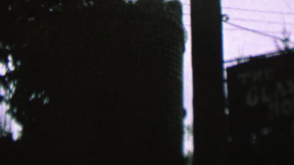 Low-exposure 8mm frame showing a shadowed wooded slope or tree mass, a nearby sign, utility pole, overhead wires, and dim sky in the supplied St. Paul clip.