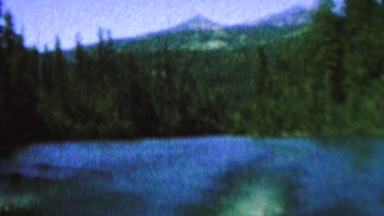 1962 8mm home-movie frame showing a blue lake, evergreen shoreline, pale sky, and distant mountain-like ridges, with no people or structures visible.