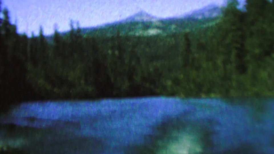 1962 8mm home-movie frame showing a blue lake, evergreen shoreline, pale sky, and distant mountain-like ridges, with no people or structures visible.
