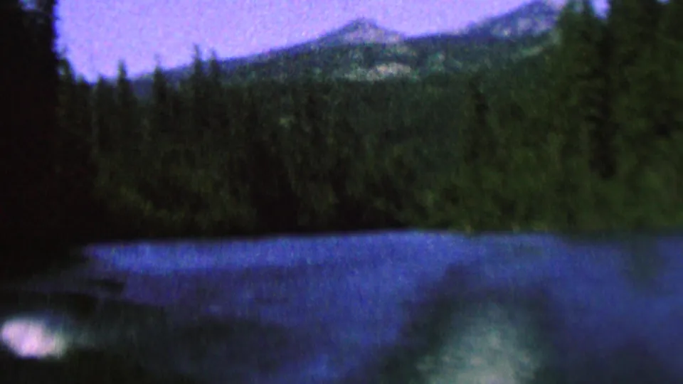 1962 8mm home-movie frame showing a forested lakeshore, blue water, distant ridge forms, and a bright reflection or wake highlight near the lower left.