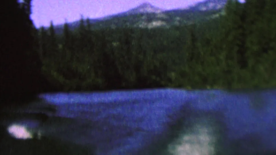 1962 8mm home-movie frame showing rippled blue lake water, evergreen trees, a dark shoreline, and distant mountain-like ridges beneath a pale sky.