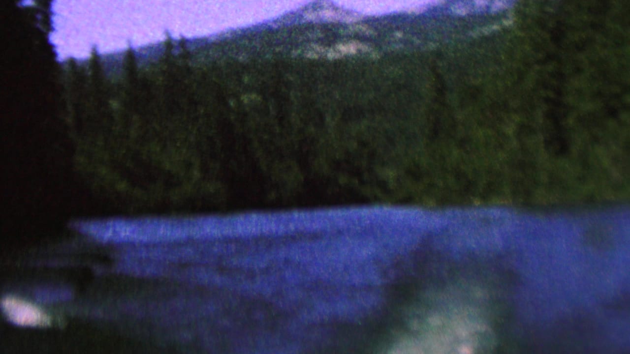 1962 8mm home-movie frame of blue river or lake water below a dark evergreen shoreline and distant mountain slopes, with a purple-tinted sky.