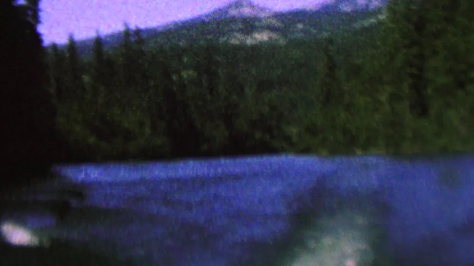1962 8mm home-movie frame of blue river or lake water below a dark evergreen shoreline and distant mountain slopes, with a purple-tinted sky.