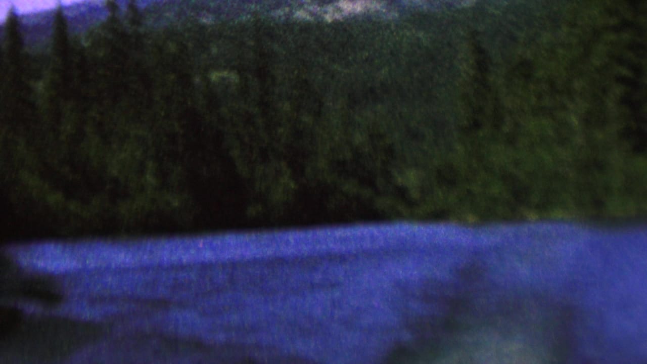 1962 8mm landscape frame showing a blue river or lake, thick evergreen trees, and a distant mountain slope under a muted sky.