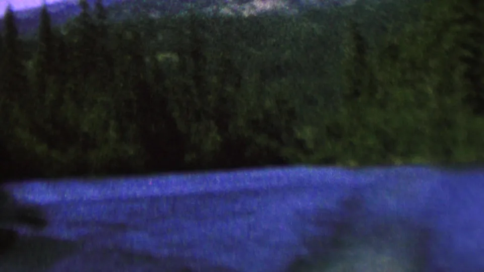 1962 8mm landscape frame showing a blue river or lake, thick evergreen trees, and a distant mountain slope under a muted sky.