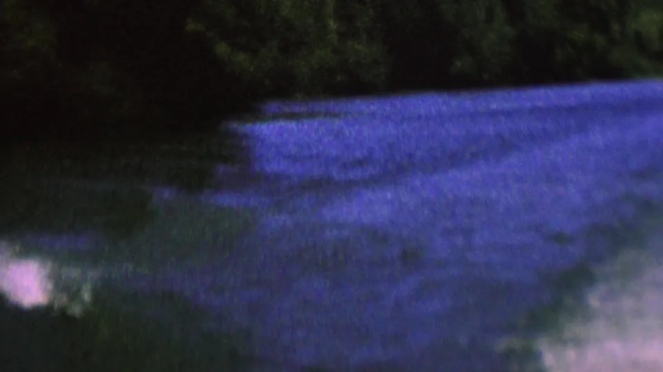 A later frame from the supplied 1962 St. Paul river clip shows the wooded bank, rippled blue-purple water, and a small pale blur near the lower edge.