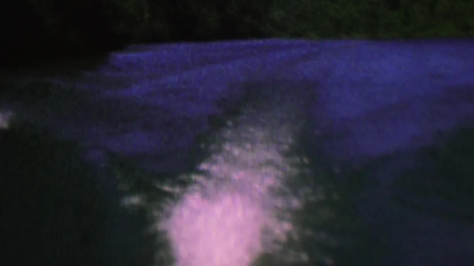 Low-light 1962 home-movie frame showing a glowing boat wake cutting through dark blue-purple water with a tree-lined shore in silhouette.