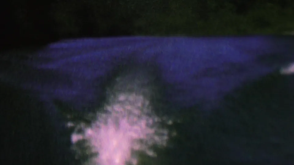 1962 8mm frame showing the boat's wake spreading across dim lake water, with soft blue-purple color and dark trees along the upper edge.