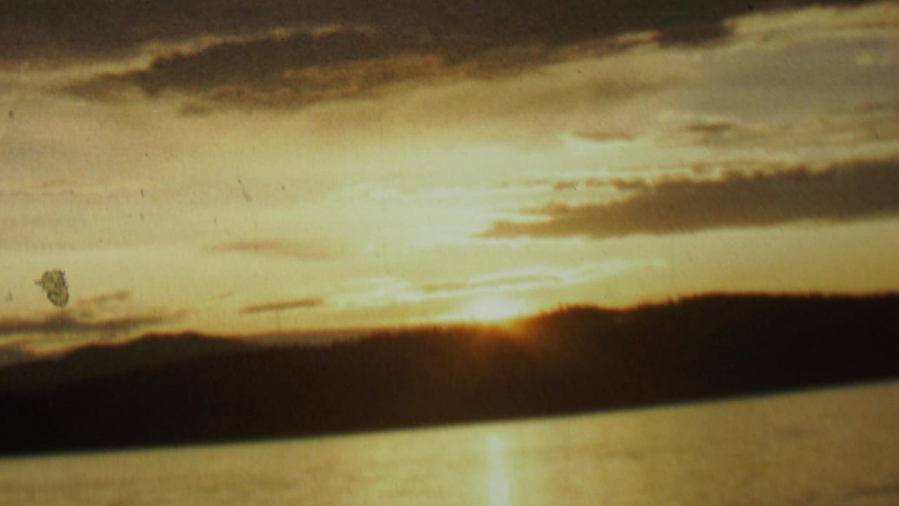 1962 8mm home-movie frame from the St. Paul, Minnesota metadata showing the sun low behind a dark shoreline, reflected across lake water beneath layered clouds.