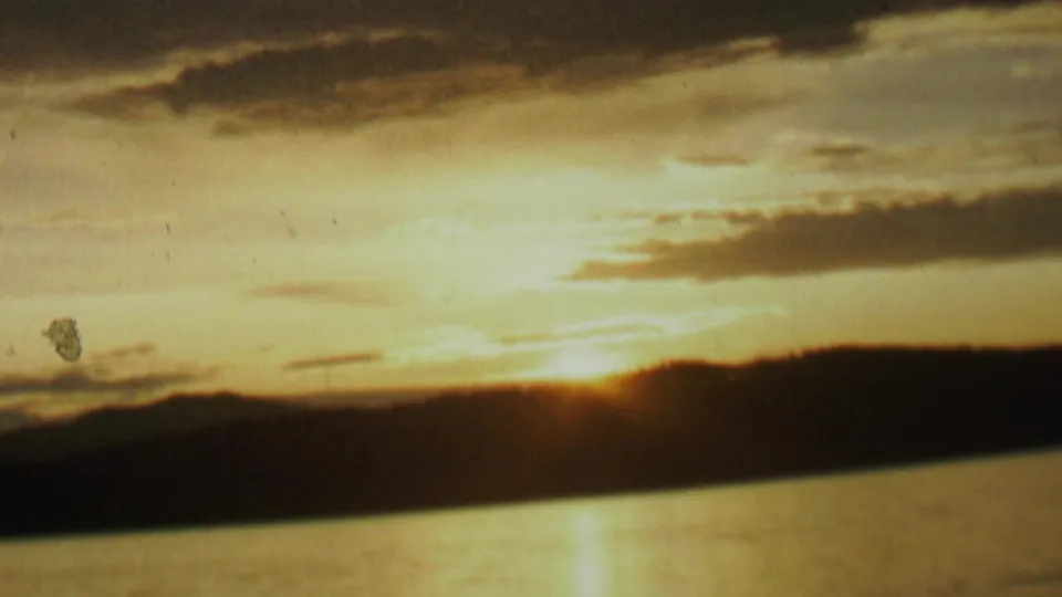 1962 8mm home-movie frame from the St. Paul, Minnesota metadata showing the sun low behind a dark shoreline, reflected across lake water beneath layered clouds.