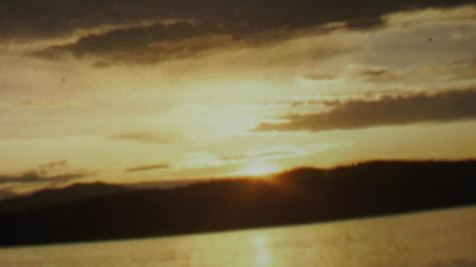 Warm 8mm frame of a low sunrise near a silhouetted lakeshore, with bright cloud cover and a narrow reflection on the water.