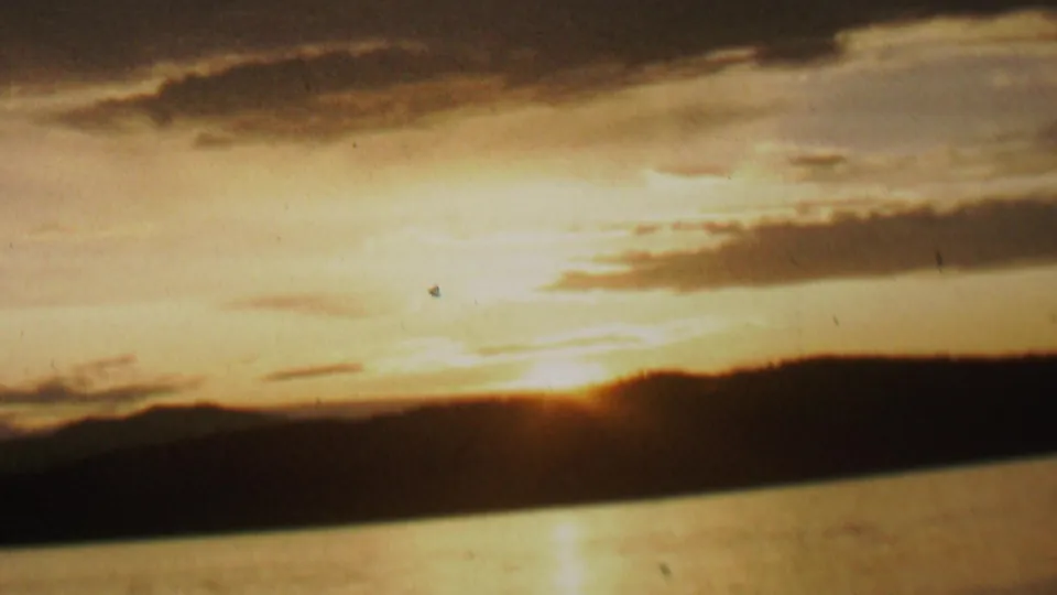 Archival 8mm frame showing a hazy sunrise over water, with a dark landform across the horizon and warm light spreading through cloud layers.