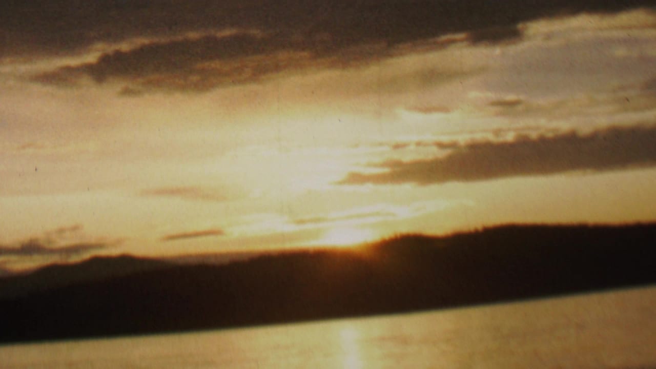 1962 8mm home-movie frame showing the sun dropping behind a dark lakeside ridge or shoreline, with amber clouds and reflected light on the water.