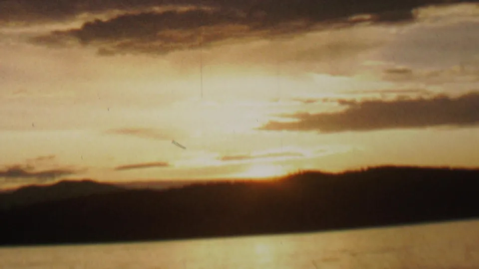 1962 8mm home-movie frame showing golden evening light over water, a shadowed shoreline or ridge, soft clouds, and visible aged-film texture.
