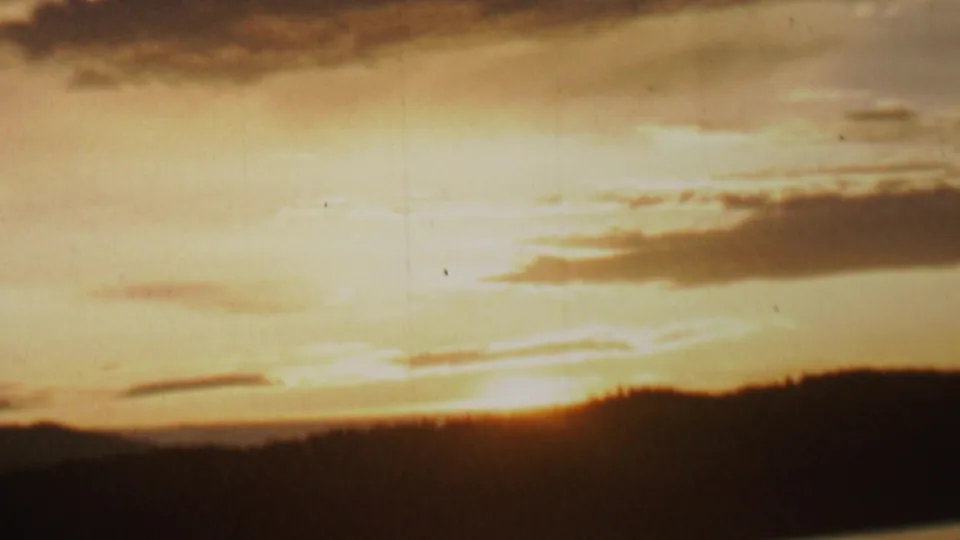 1962 8mm home-movie frame showing the sun partly hidden behind a dark lakeside ridge or shoreline, with warm cloud layers across the sky.