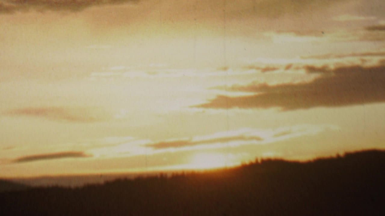 Warm 1962 8mm home-movie frame from the supplied Minnesota clip showing the sun setting behind a dark wooded ridge under streaked clouds.