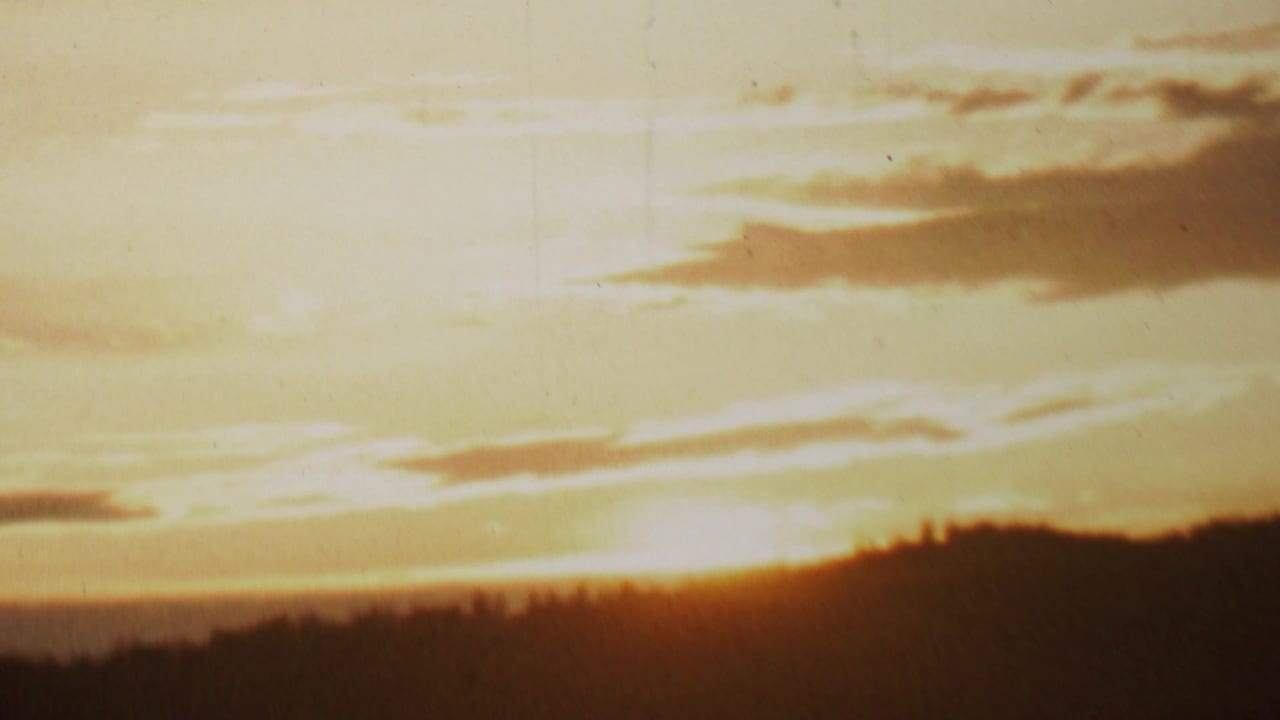 Warm 8mm frame showing a low orange sun near a silhouetted treeline, with soft clouds and visible film texture from the supplied 1962 lakeside footage.