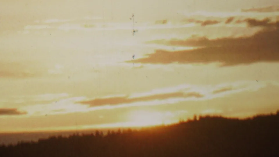 Overexposed golden sunset sky in the supplied 1962 8mm lake scene, with cloud bands above a dark wooded horizon and visible vertical film wear.