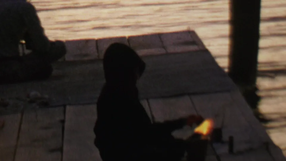 1962 8mm lakeside frame from the supplied Minnesota clip showing a young man or seated figure in silhouette on a dock, with reflective water and a small flame nearby.