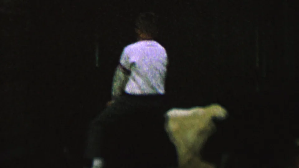 A small rider in a light shirt sits on the cow as the low-light 8mm frame shows more black space and faint background structure around the figures.
