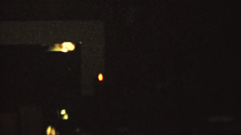 Grainy 1962 home-movie frame showing a mostly black interior, a bright warm reflection near the lower left, and a few isolated candlelike glows.