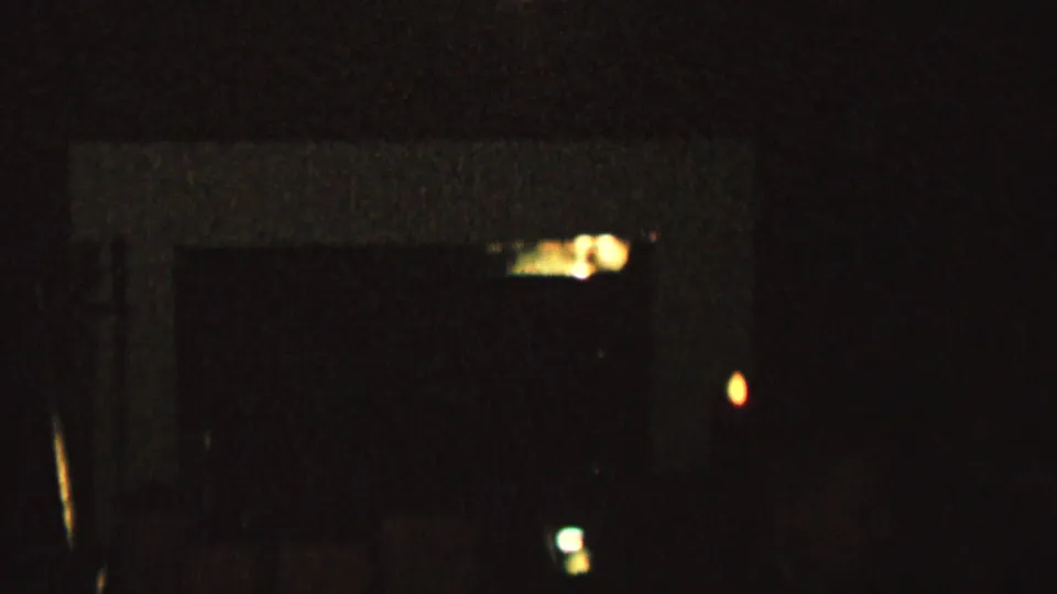 Low-exposure 8mm frame with a pale horizontal interior shape, small orange lights, faint reflections, and no clearly identifiable people or objects.