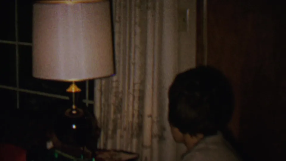 In a 1962 St. Paul home-movie frame, a woman with short dark hair is seen from behind near a beige-shaded lamp, cream curtains, and a dark nighttime window.