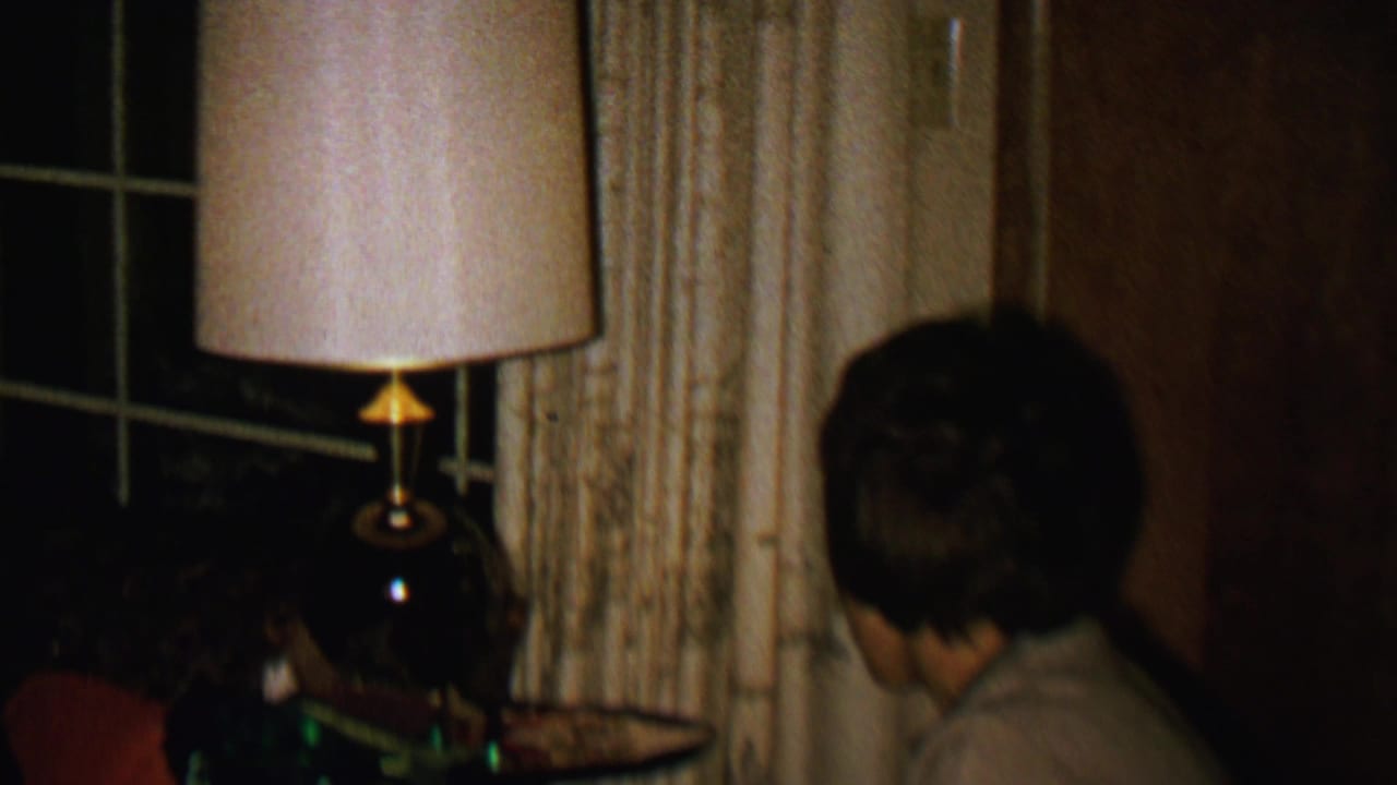 The frame shows the woman turned away in a dim 1962 domestic interior, with a lit table lamp, cream curtains, a dark window, and wood paneling visible around her.