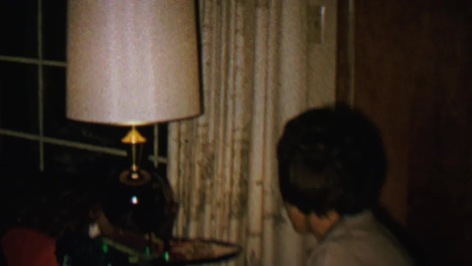 The frame shows the woman turned away in a dim 1962 domestic interior, with a lit table lamp, cream curtains, a dark window, and wood paneling visible around her.